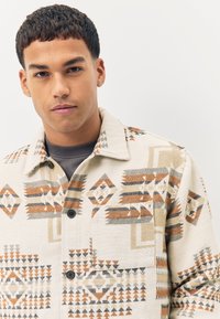 Chemise à motif de couleur claire présentant des dessins géométriques en marron, gris et noir, avec un col, des boutons et une poche poitrine.