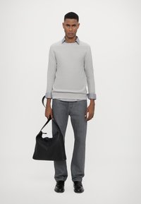 Hellgrauer gesteppter Sweater, verblichene graue Jeans und schwarze Lederhandtasche. Schwarze Schuhe runden den Look ab. Das Model steht vor einem weißen Hintergrund.