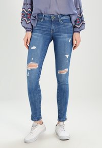 Jeans ajustados de denim azul medio con áreas desgastadas y dobladillos deshilachados. Combinados con zapatillas blancas. Top suelto de manga larga con bordados.
