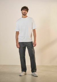 Camiseta blanca de manga corta combinada con pantalones cargo gris oscuro. Modelo con zapatillas blancas se encuentra frente a un fondo neutro.