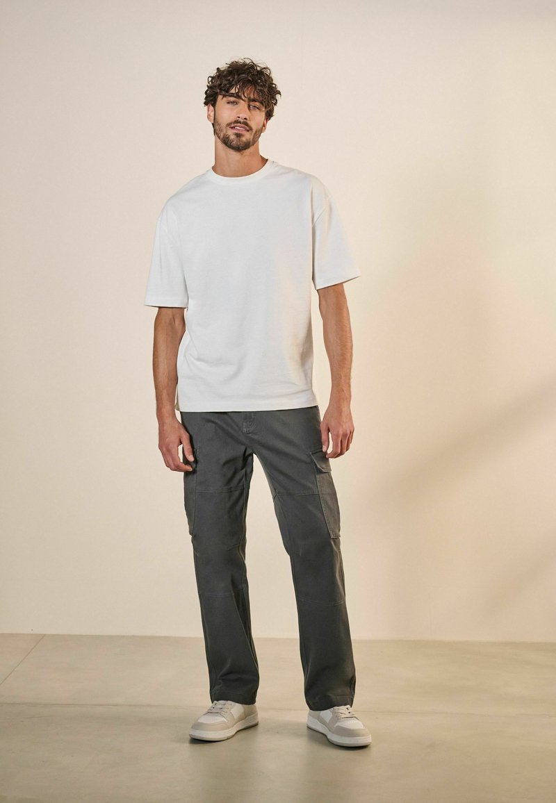 Camiseta blanca de manga corta combinada con pantalones cargo gris oscuro. Modelo con zapatillas blancas se encuentra frente a un fondo neutro.