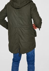 Olivgrön parkasjacka med huva, dragsko i midjan och åtsittande nederkant. Har en ledig passform och strukturerat tyg. Bakifrån visad.