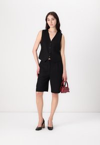 Svart ärmlös väst med rundad nederkant, framknappar och matchande svarta skräddarsydda shorts. Tillbehören inkluderar en liten röd handväska och svarta klackar.