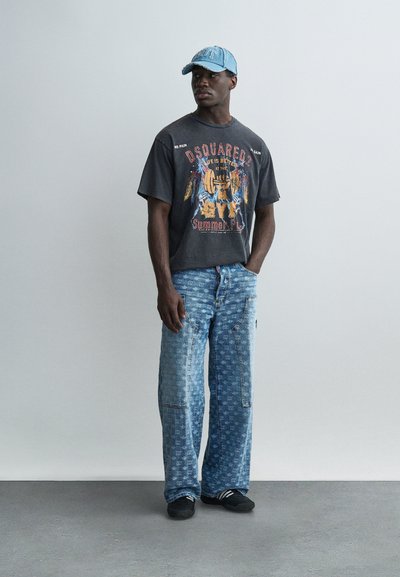 Άνδρας που φοράει μαύρο T-shirt με σχέδιο, ανοιχτό μπλε τζιν με σχέδια, μαύρα αθλητικά παπούτσια και ανοιχτό μπλε καπέλο, στέκεται μπροστά σε έναν απλό γκρι τοίχο.