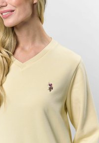 Dzeltena V-veida jaku ar ribotu apkakli un garām piedurknēm, ar nelielu izšūtu logo uz krūšu. Mīksts kokvilnas audums.