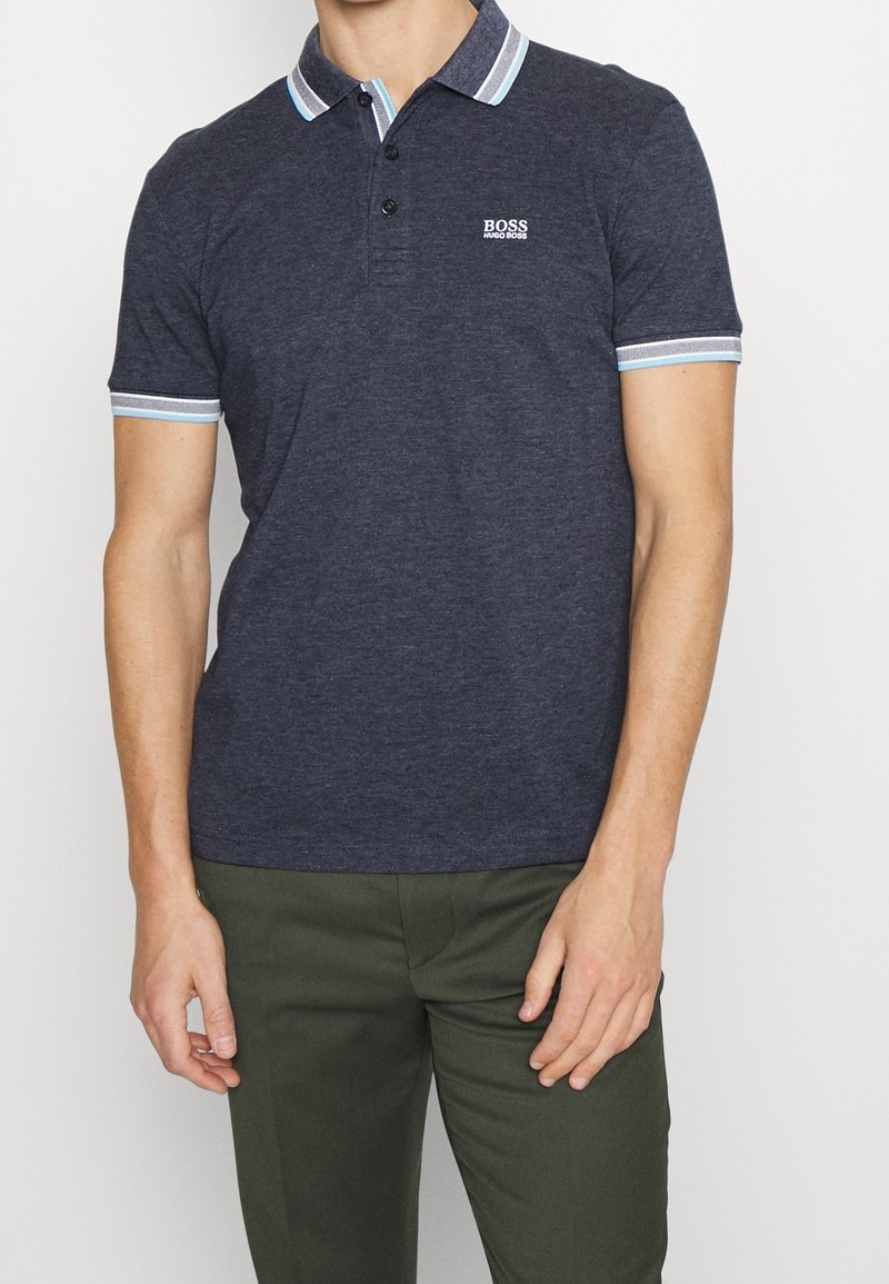 Homme portant un polo bleu marine Hugo Boss avec col et poignets rayés, assorti d’un pantalon vert olive.