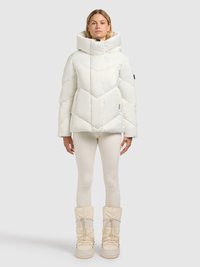 Veste matelassée blanche avec un col montant, motif matelassé en losanges, et un logo sur la manche. Associée à des leggings beige et des bottes montantes jusqu'au genou.
