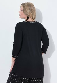 Schwarzes Tunika-Oberteil mit Dreiviertelärmeln, das mit Perlenakzenten an den Ärmeln und am Ausschnitt verziert ist. Weicher Stoff, leicht verlängertem Saum.