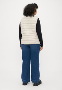 Šviesiai smėlio spalvos pūkinė liemenė su horizontaliomis siuvimo linijomis, dėvima ant juodos ilgomis rankovėmis marškinėlių. Suporuota su plačiomis mėlyno džinsinio audinio kelnėmis ir juodais batais.