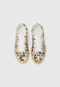 Brune og hvite prikkete sneakers med en teksturert overdel, hvite snørebånd og en lys gummisåle. Merking er synlig inni skoen.
