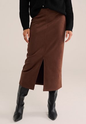 Femme portant une jupe midi marron avec une fente à l'avant, un haut noir à manches longues et des bottes noires hautes jusqu'au genou, debout devant un fond neutre.