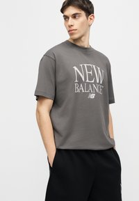 Mladý muž v šedej čiernej tričku New Balance a čiernych nohaviciach, stojaci s rukami v vreckách, pozerajúci sa do strany na jednoduchom pozadí.