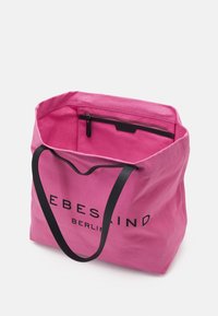 Różowa torba tote z płótna z czarnymi uchwytami, z wewnętrzną kieszenią na zamek i nadrukowanym logo z przodu. Miękka tekstura, przestronny design.