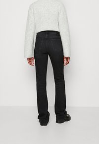 Henkilö, joka on pukeutunut mustiin suoriin farkkuihin, mustiin kenkiin ja vaaleanharmaaseen neuleeseen, seisoo selin yksinkertaista valkoista taustaa vasten.