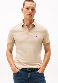Béžové polokošile z hladké tkaniny, s klasickým límcem, krátkými rukávy a malým logem na levé straně hrudi. Nošeno s modrými džínami.