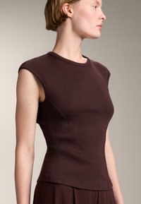 Mörkbrun, ribbad ärmlös topp med rund halsringning och figurnära design. Har subtila sömdetaljer och en slät textur.