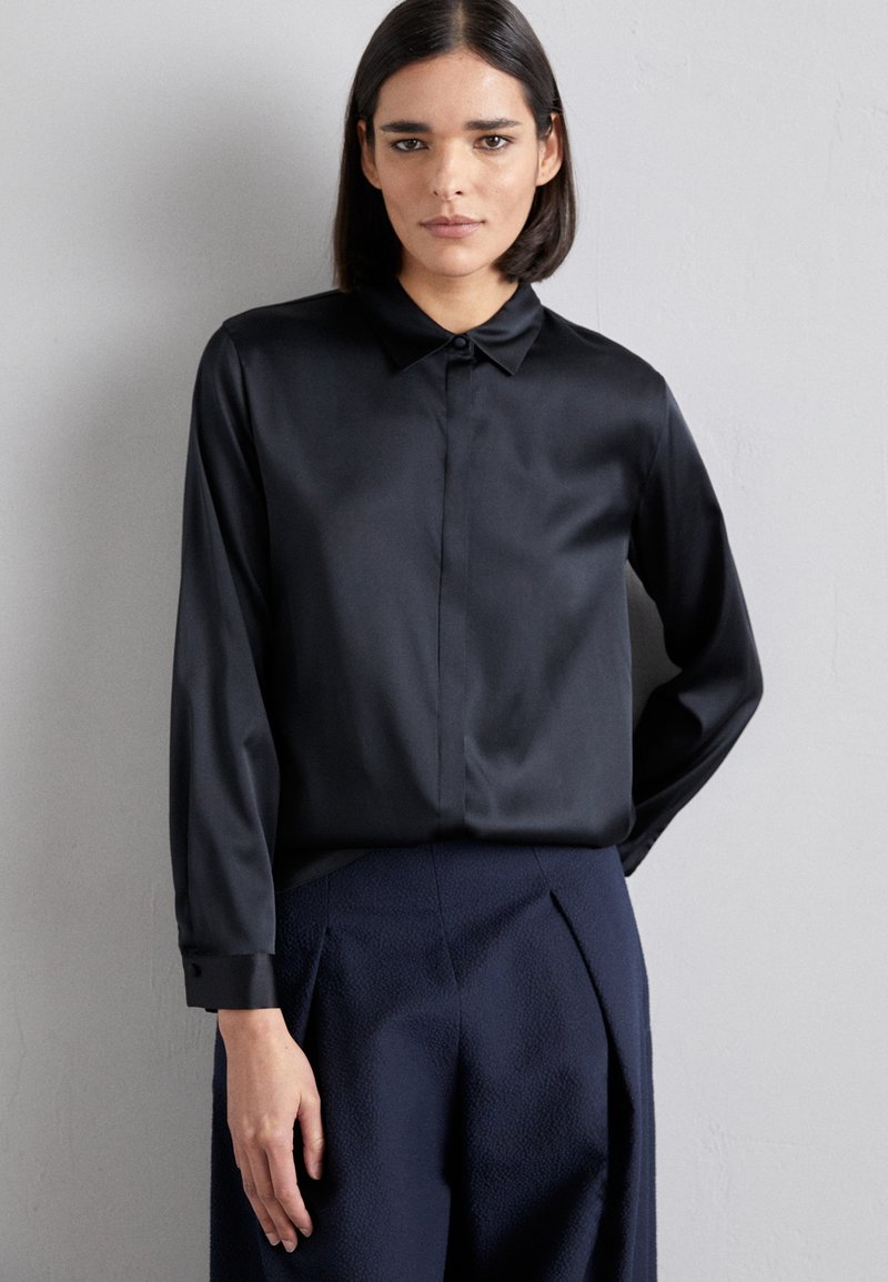 Chemise longue noire en satin à manches longues avec un col classique, coupe droite et poignets boutonnés. Portée avec un pantalon texturé bleu marine.