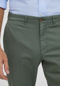 Olivově zelené chino kalhoty s hladkou texturou, vybavené předním zipem, zapínáním na knoflík a bočními kapsami. Kombinovány se světle modrou košilí.