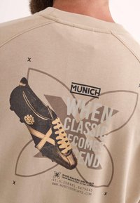 Sudadera beige con un gráfico de estilo retro que presenta una zapatilla negra y amarilla, el texto "MUNICH" y "CUANDO LO CLÁSICO SE CONVIERTE EN TENDENCIA."