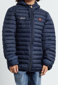 Marinblå puffjacka med quiltat mönster, med dragkedjestängning och huva. Varumärkeslogotyp på bröstet och distinkt accentdetaljering.