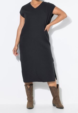 Personne portant une robe midi noire à manches courtes avec une fente latérale et des bottes de cowboy brodées marron, posant avec une main sur la hanche.