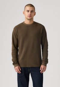 Καφέ μπλούζα με μακριά μανίκια και ριπ υφή, με στρογγυλή λαιμόκοψη, υφασμάτινη υφή και εφαρμοστά μανσέτες, συνδυασμένη με σκούρα μπλε τζιν παντελόνια.