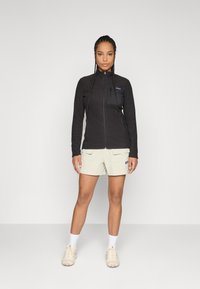 Vrouw met gevlochten haar in een zwart ritssluitingjack, beige cargoshorts, witte sokken en beige sneakers, staand tegen een effen achtergrond.