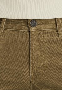 Pantaloni in velluto a coste marrone con una finitura testurizzata, chiusura con bottone superiore e un dettaglio tasca sul lato sinistro.