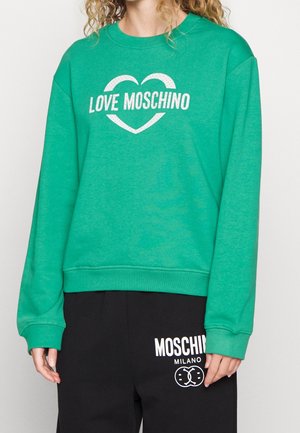 Personne portant un sweat-shirt vert avec « LOVE MOSCHINO » et un cœur, associé à un pantalon noir arborant le logo blanc Moschino Milano.