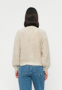 Person med skulderlangt mørkt hår iført en kremfarget strikkegenser og blå jeans, vendt bort fra kameraet mot en ensfarget bakgrunn.