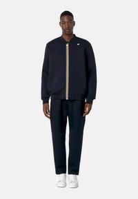 Felpa blu navy con zip, colletto e polsini a coste, caratterizzata da una striscia verticale dorata e rossa. Abbinata a pantaloni joggers blu navy e sneakers bianche.