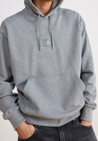 Grå huvtröja i mjukt tyg, med en framficka, justerbara snoddar och en logotyp patch på bröstet. Ribbad mudd vid ärmar och fåll.