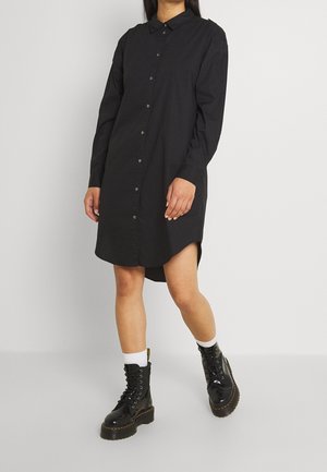 Persoon draagt een zwart, lang mouw, knoopjurk gecombineerd met witte sokken en zwarte veterplateauhakken, lopend tegen een effen achtergrond.