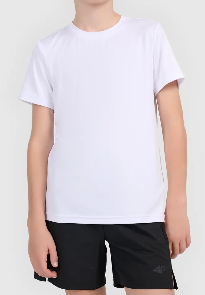 Personne portant un t-shirt blanc uni à manches courtes et un short noir, debout devant un fond clair uni.