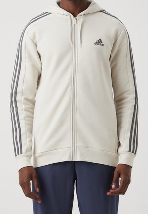 Homme portant un sweat à capuche zippé blanc Adidas avec des rayures grises sur les manches et un pantalon sombre sur un fond uni.