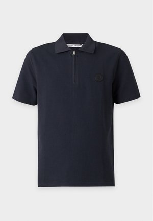 Mężczyzna ubrany w czarną koszulkę polo z krótkim rękawem, szare luźne spodnie i beżowe sneakersy stoi na tle jednolitego jasnego tła.