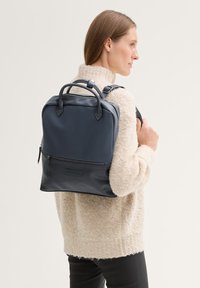 Zaino in pelle blu navy con due manici superiori, spallacci regolabili e tasca frontale con zip. Texture liscia con linee pulite e design minimalista.