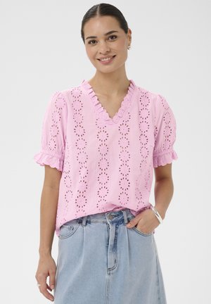 Femme souriante, portant un chemisier rose à manches courtes en broderie anglaise avec des bords volantés et une jupe en denim bleu clair, la main dans la poche de la jupe.