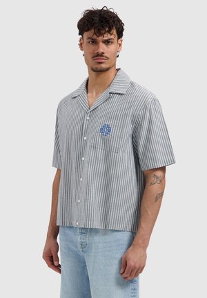 Man wearing a gray and white vertical striped short-sleeve button-up shirt with light blue jeans and visible arm tattoos.