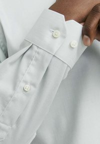 Camicia grigia chiara in tessuto texturizzato, con dettagli a bottone sui polsini. Design del colletto semplice e classico con cuciture visibili.