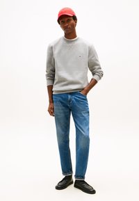 Sudadera gris con cuello redondo, vaqueros azules y zapatos negros. Una gorra roja completa el atuendo, añadiendo un toque de color.