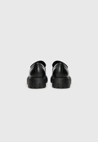 Black leather shoes with a textured surface, featuring a chunky rubber sole and rounded heel, designed without traditional laces.
