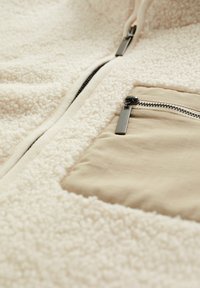 Beige plüss pulóver, bolyhos textúrával, elöl cipzárral és egy kontrasztos zsebbel, amelyen sima anyagú elegáns cipzár található.