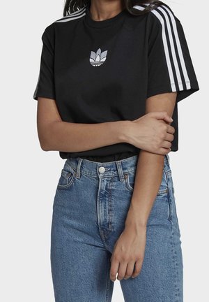 Persoon draagt een zwart Adidas t-shirt met witte strepen op de mouwen en een logo op de borst, gecombineerd met een hoog getailleerde blauwe spijkerbroek.