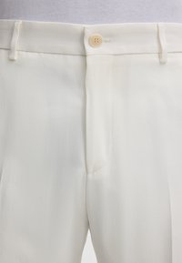 Beige Hose mit einem Knopf und Gürtelschlaufen, von der Taille bis zur Mitte des Oberschenkels vor einem weißen Hintergrund gezeigt.