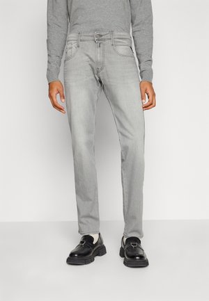 Homme portant un jean gris clair, des mocassins en cuir noirs et un haut gris à manches longues, debout devant un fond blanc uni.