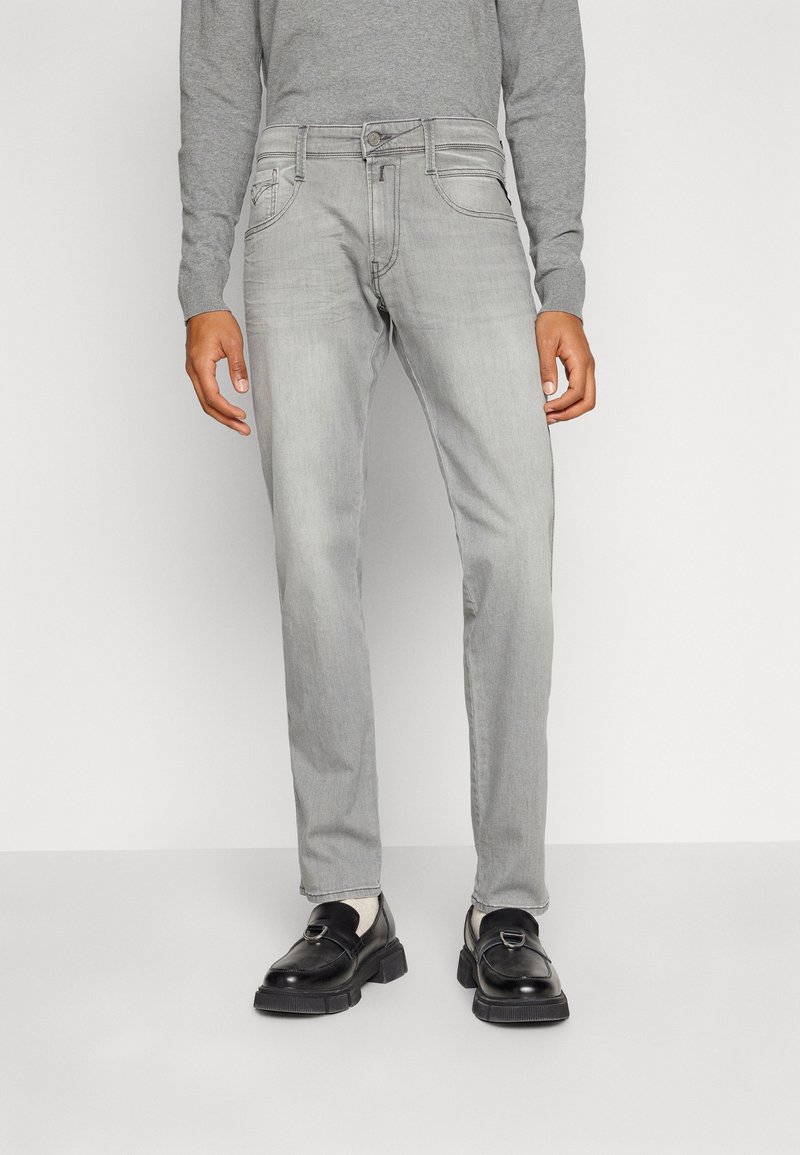 Homme portant un jean gris clair, des mocassins en cuir noirs et un haut gris à manches longues, debout devant un fond blanc uni.