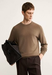 Brauner gestrickter Pullover mit Rundhalsausschnitt, kombiniert mit dunkelhimmelbraunen maßgeschneiderten Hosen. Die Person hält eine schwarze Wildledertasche mit kontrastierender Einfassung.