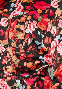 Tissu noir à motifs floraux avec de grandes fleurs rouges, roses, orange, vertes et bleues, comprenant des boutons noirs et une poche avec un rabat à bouton.