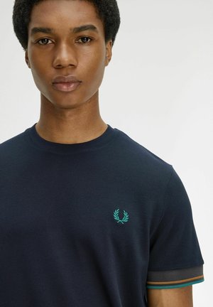 Young person with short curly hair wearing a navy blue t-shirt with teal laurel logo and striped sleeve cuffs against a plain background.